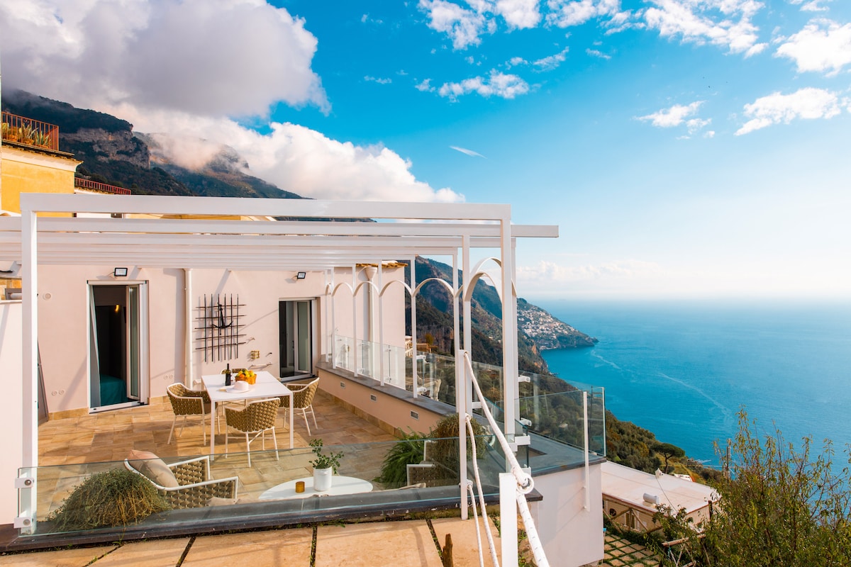 An outdoor terrace offers a panoramic view of the sea and the surrounding landscape. Comfortable seating arrangements are positioned beneath a stylish pergola, while glass balustrades provide an unobstructed sense of openness. The bright blue sky complements the serene waters below.