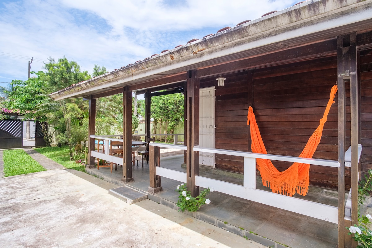 A wooden structure features a porch area with a vibrant orange hammock draped between two support beams. A spacious outdoor dining table sits nearby, surrounded by greenery and well-maintained grass. The tranquil setting is complemented by the natural light filtering through the surrounding trees.