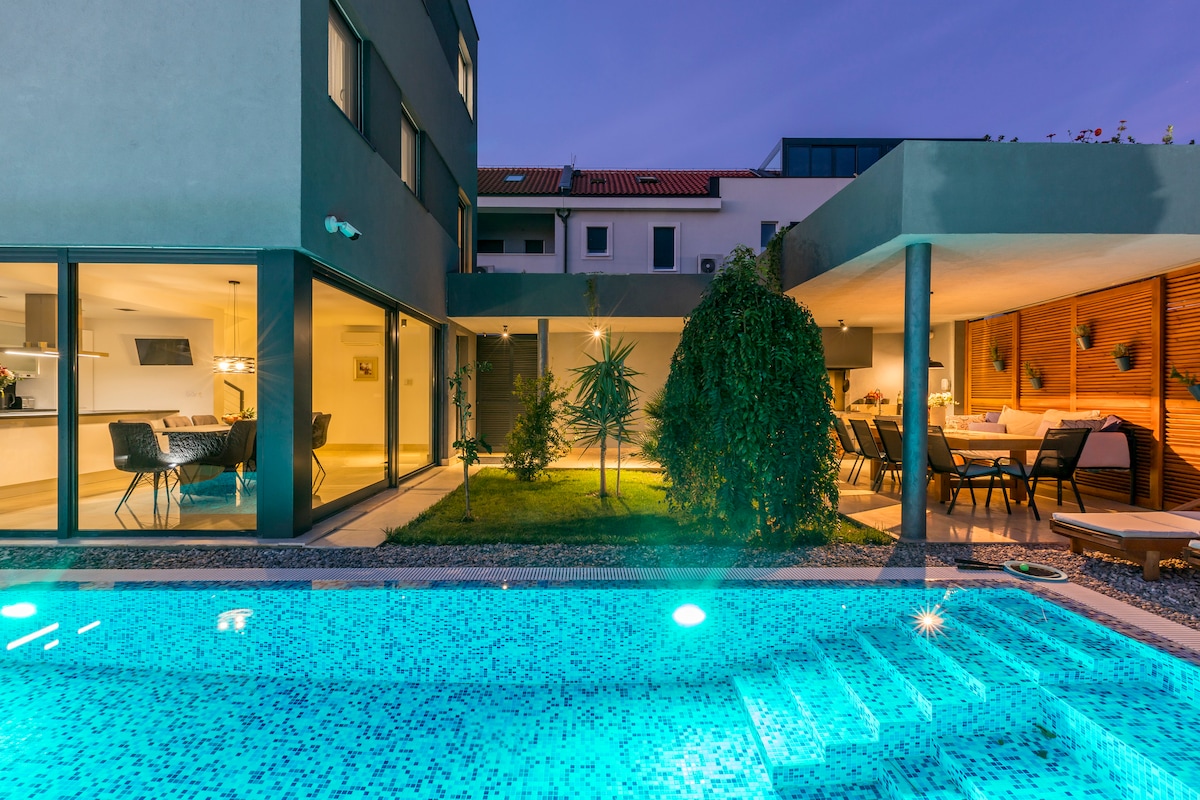 A modern villa exterior is highlighted at dusk, with a sparkling turquoise pool in the foreground. Large glass doors create a seamless transition between indoor and outdoor spaces. The well-manicured garden area, illuminated softly, includes a cozy dining space adjacent to the pool.