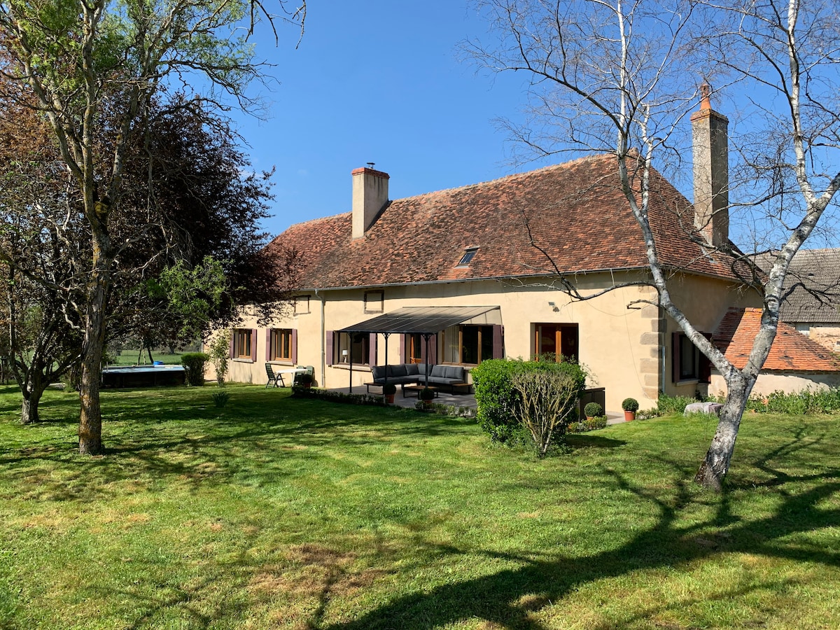A spacious country house is set on a grassy lawn, framed by trees. Large windows provide natural light to the interior, while a covered terrace offers outdoor seating. A pool can be seen in the background under clear blue skies.