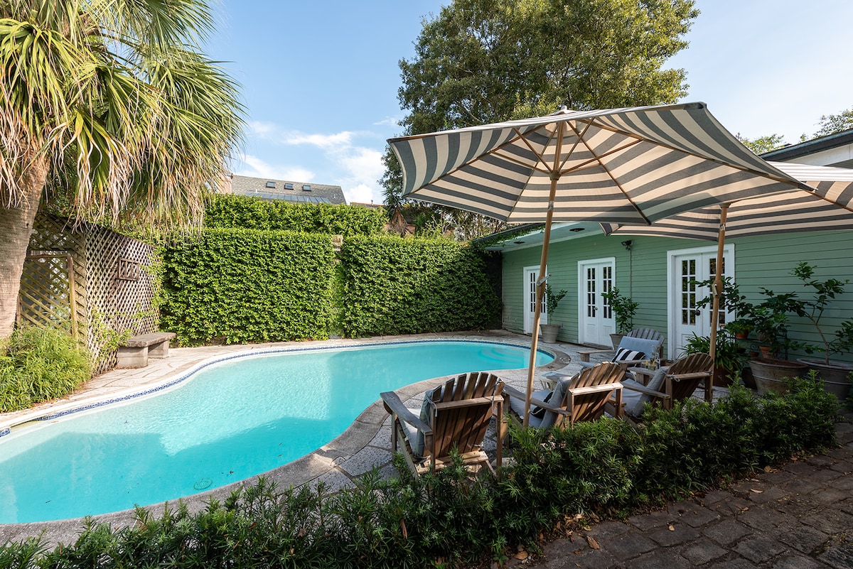 An inviting pool area is framed by lush greenery and a vibrant blue sky. Lounge chairs are positioned by the water's edge under a large striped awning, offering a shaded spot to relax. The pool reflects natural light, enhancing the calm ambiance of the courtyard.