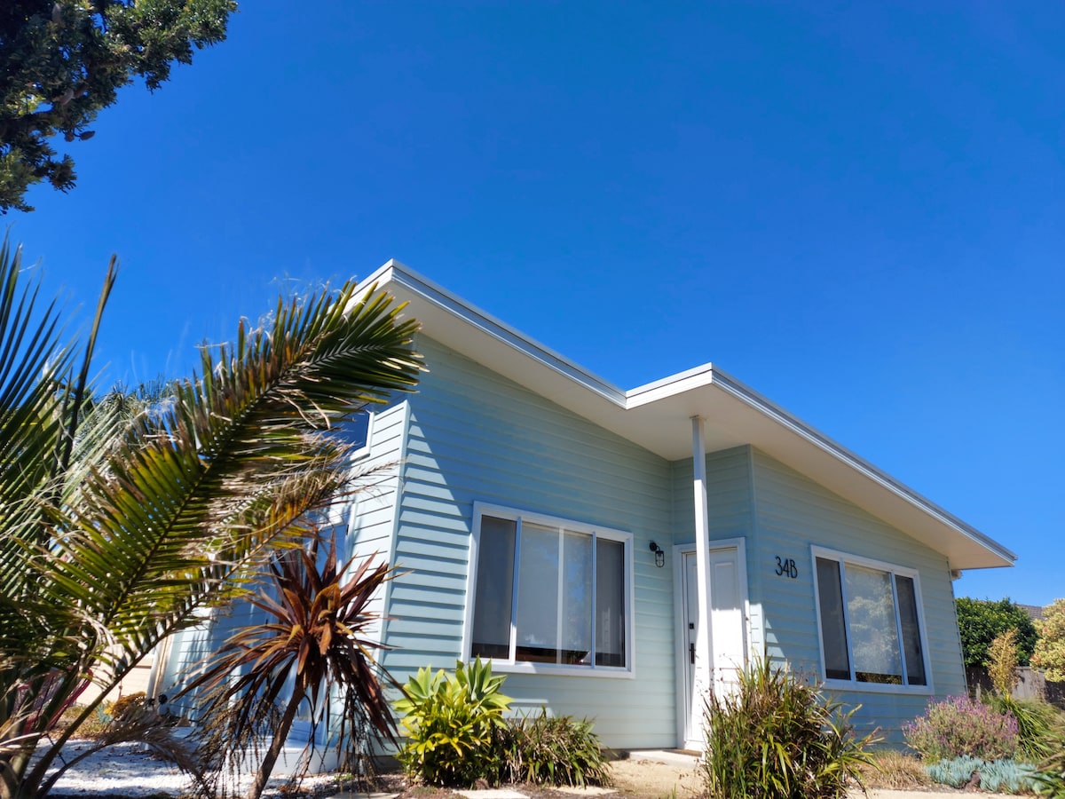 A modern guest house with light blue cladding is shown under a clear blue sky. Lush greenery and palms surround the exterior, highlighting the inviting entrance with a white door and large windows that provide natural light to the interior.