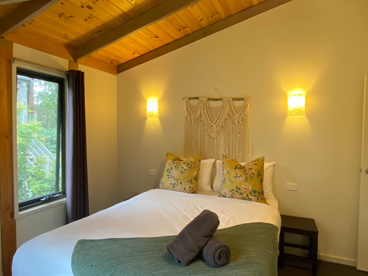 A bright sleeping area is highlighted by a spacious bed dressed with decorative pillows and a cozy blanket. Natural light enters through a window adorned with dark curtains, complemented by warm wall lighting and wooden ceiling beams.