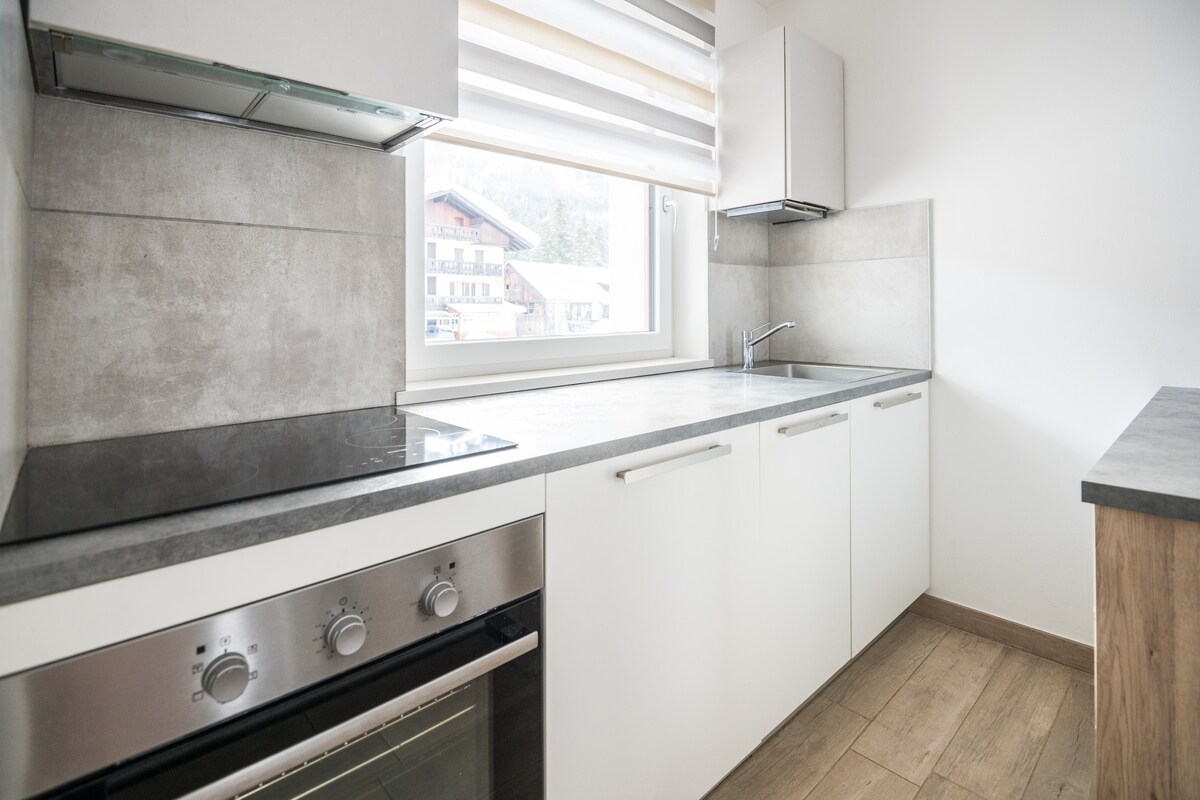 A modern kitchen is presented with sleek cabinetry and a built-in oven. A large window allows natural light to illuminate the space, showcasing a countertop with a minimalist design. The walls are light-colored, enhancing the overall clean and functional aesthetic.