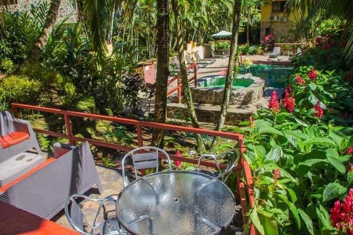 A view from the private terrace presents a vibrant garden filled with lush greenery and colorful flowers. The shared pool and whirlpool are visible in the background, surrounded by tropical plants, while outdoor seating is arranged around a glass table nearby.