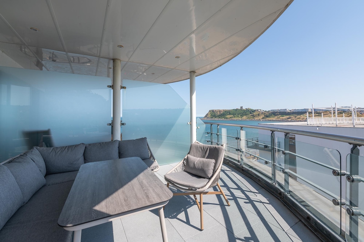 A private wrap-around balcony features modern outdoor seating, including a cozy sofa and a stylish armchair. Expansive glass panels and a sleek table provide unobstructed views of the serene surroundings, enhancing the outdoor experience.
