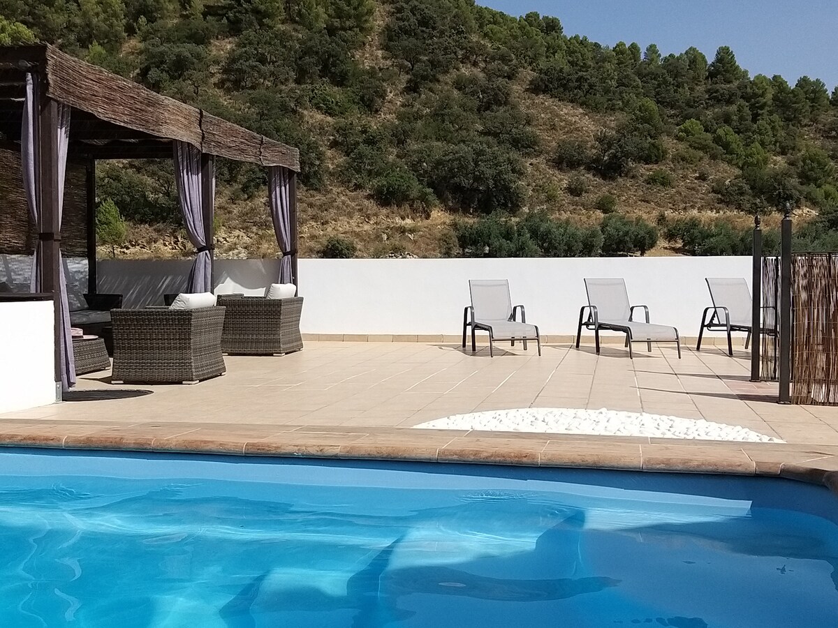 A spacious terrace features a welcoming heated pool surrounded by sun loungers and a shaded lounge area with comfortable wicker seating. The backdrop presents natural greenery, enhancing the relaxing atmosphere.