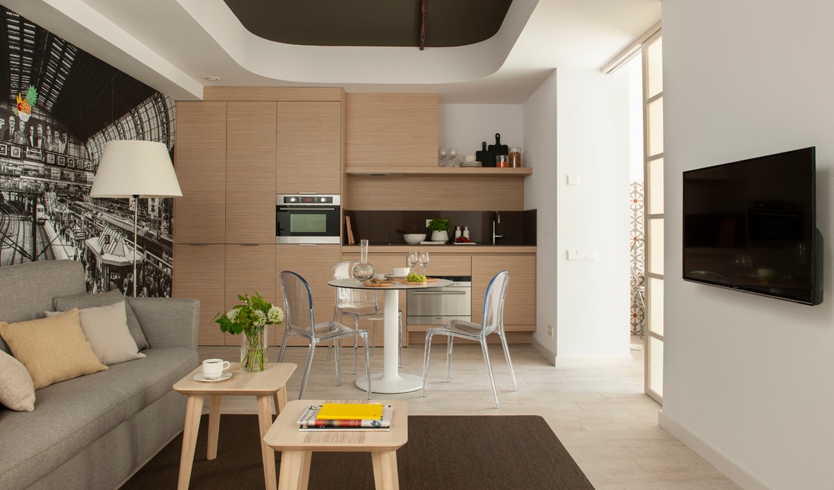 A modern living and dining area features a sleek, fully equipped kitchen with minimalist cabinetry. A round dining table is set for two, accompanied by transparent chairs. A comfortable sofa in soft grey is complemented by decorative cushions, while a flat-screen TV is mounted on the wall.