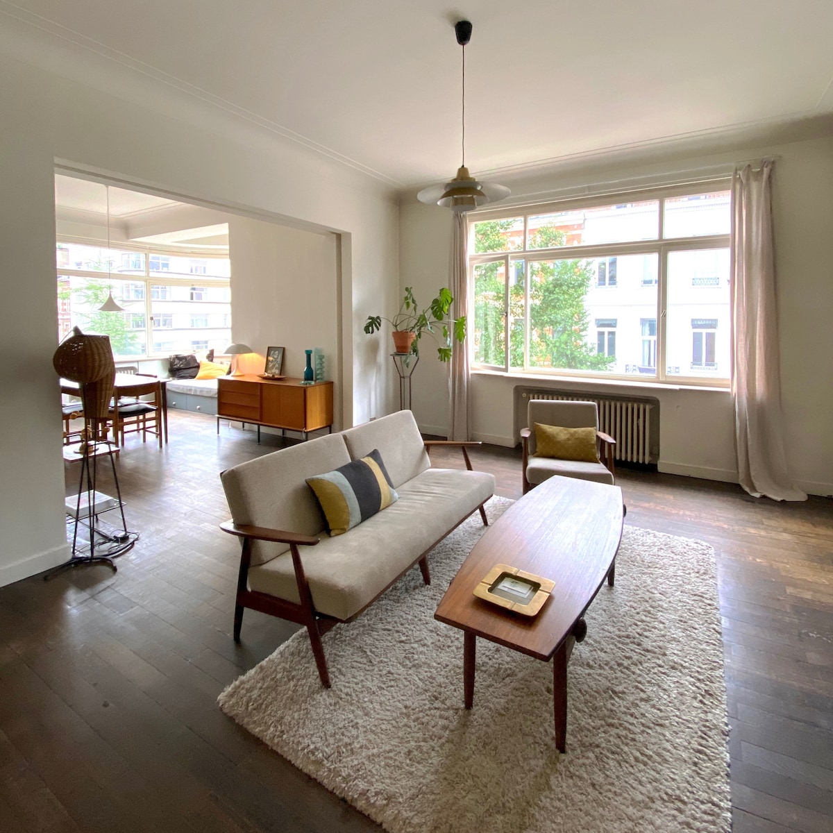 A spacious living area is displayed, featuring a comfortable sofa and a wooden coffee table. Light filters through large windows, illuminating the dark wooden flooring. A dining area is visible through an open entry, with a stylish cabinet and plants enhancing the atmosphere.