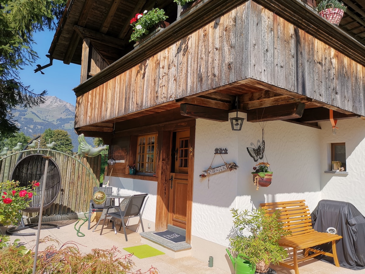 The entrance of the chalet is framed by natural wooden walls and a decorated balcony featuring potted flowers. A small seating area with a bench and table is visible, along with a barbecue grill. Mountain views can be seen in the background.