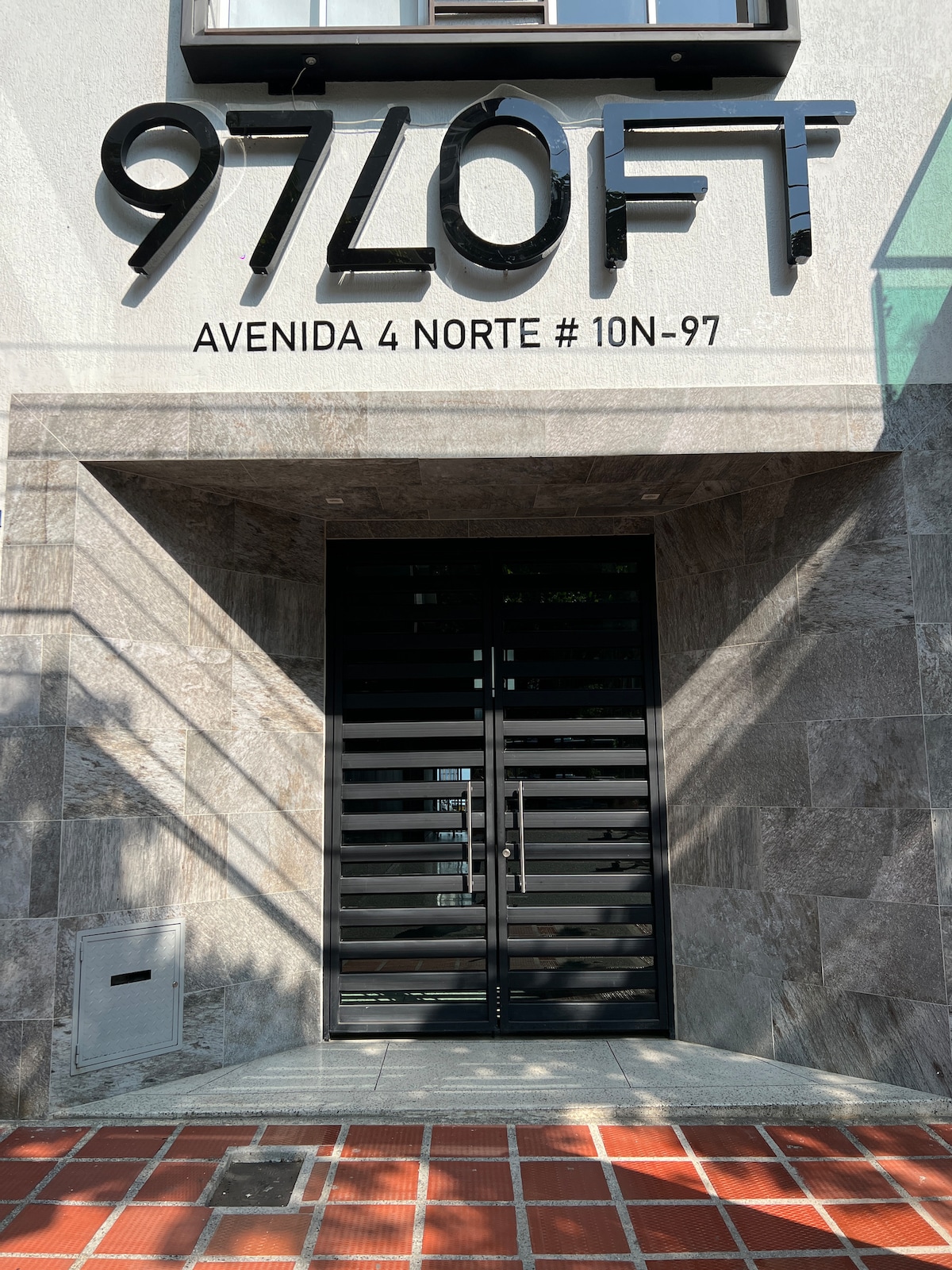 The entrance to the building is showcased, featuring a modern design with a sleek black door. Above the door, the building name '97 LOFT' is displayed prominently. The surrounding architecture includes stone accents and a tiled walkway, inviting guests into a contemporary space.