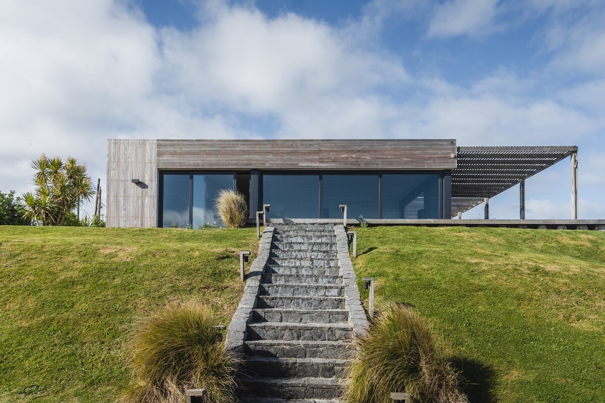The exterior of the house is showcased from a stone pathway lined with green grass. Large glass windows dominate the front, and a shaded patio extends outward, providing a welcoming space for outdoor relaxation.