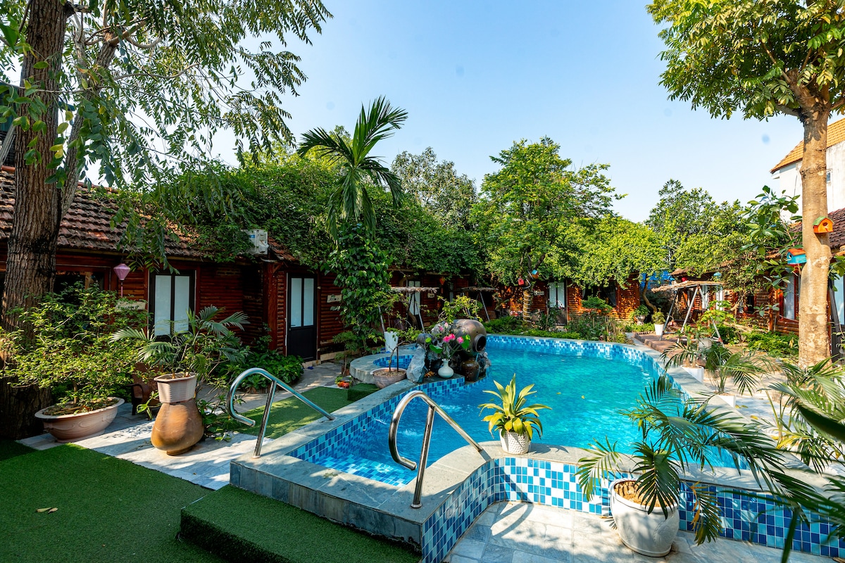 A swimming pool is centrally located within a lush garden, surrounded by greenery and rustic bungalows. The pool’s inviting blue tiles are complemented by vibrant landscaping and palm trees, creating a serene outdoor atmosphere ideal for relaxation.