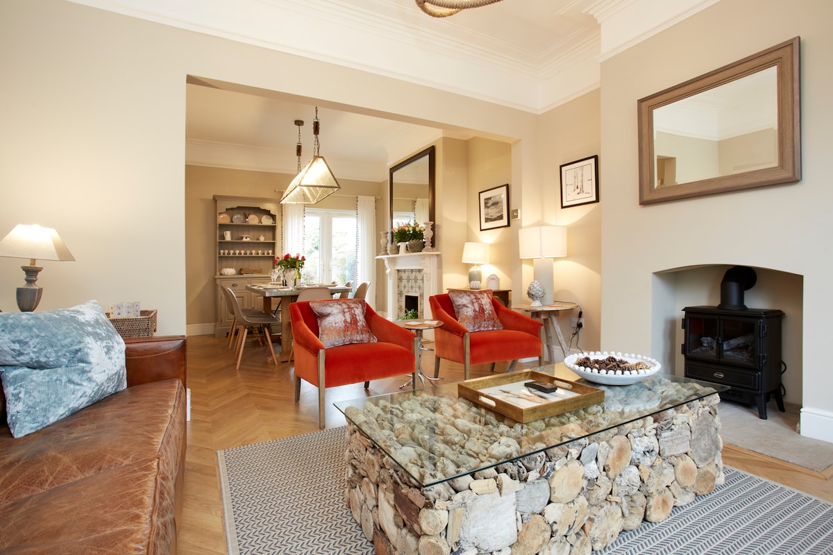A welcoming living area features a rich leather sofa and striking orange armchairs. A unique coffee table composed of stone is positioned on a patterned rug. Natural light enters through expansive windows, illuminating the neutral tones and stylish decor, creating an inviting gathering space.