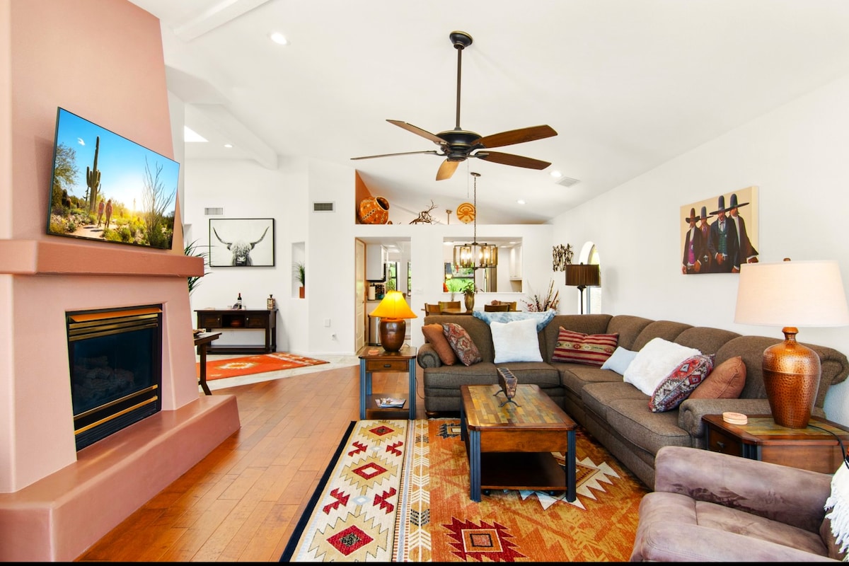 A spacious living area features a sectional sofa with decorative pillows surrounding a wooden coffee table. A fireplace is situated below a flat-screen TV, with vibrant area rugs adding color to the hardwood floors. Natural light filters in through large windows, enhancing the open feel.