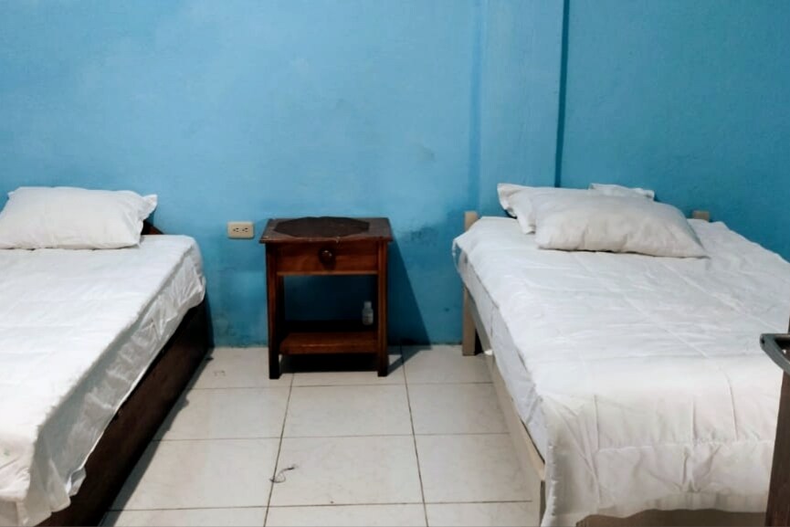 A room features two neatly arranged beds with white linens, positioned against a blue wall. A small wooden nightstand is located between the beds, providing a convenient surface for personal items. The floor is covered with light-colored tiles.