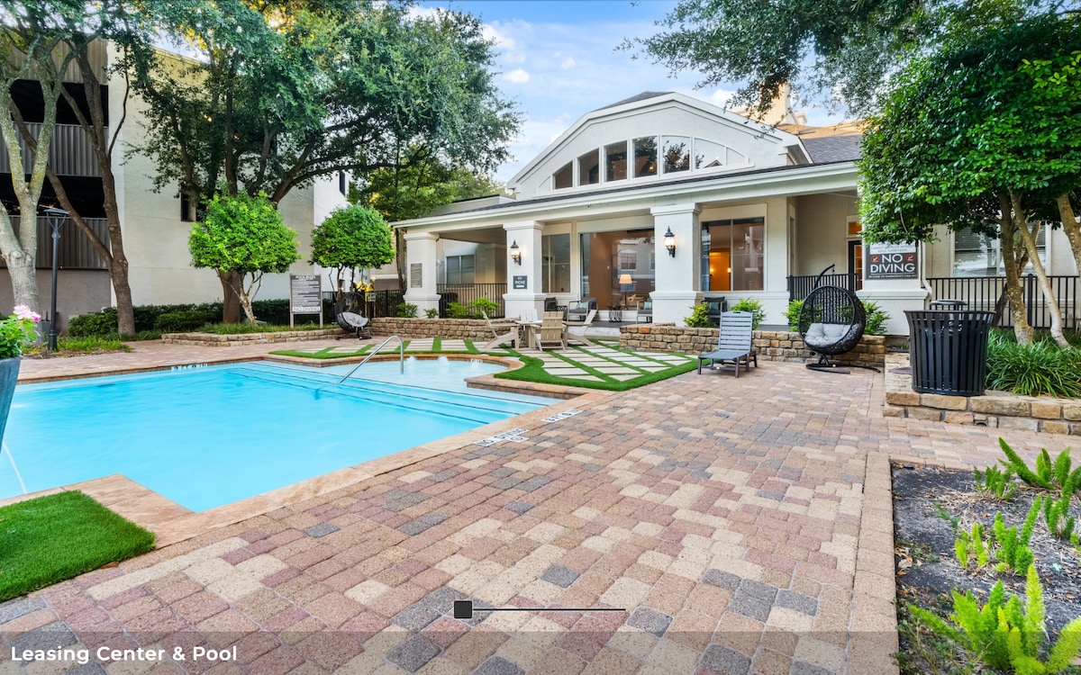 A well-maintained pool area features a clear water pool surrounded by colorful pavers and landscaped borders. Lounge chairs are positioned near the pool, while a welcoming leasing center is visible in the background, framed by lush greenery and outdoor seating.