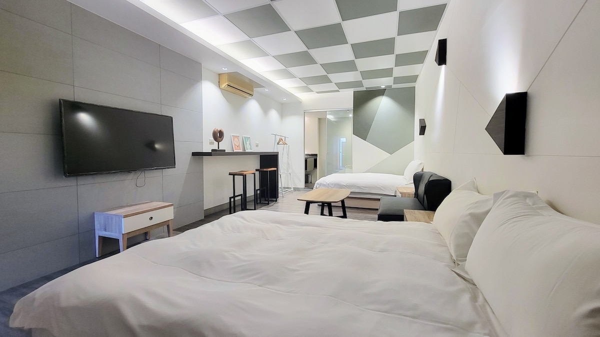 A modern room featuring two spacious beds and a minimalist design. A large flat-screen TV is mounted on the wall, while a kitchenette area with bar seating is visible in the background. Neutral colors and geometric patterns are present on the walls and ceiling.