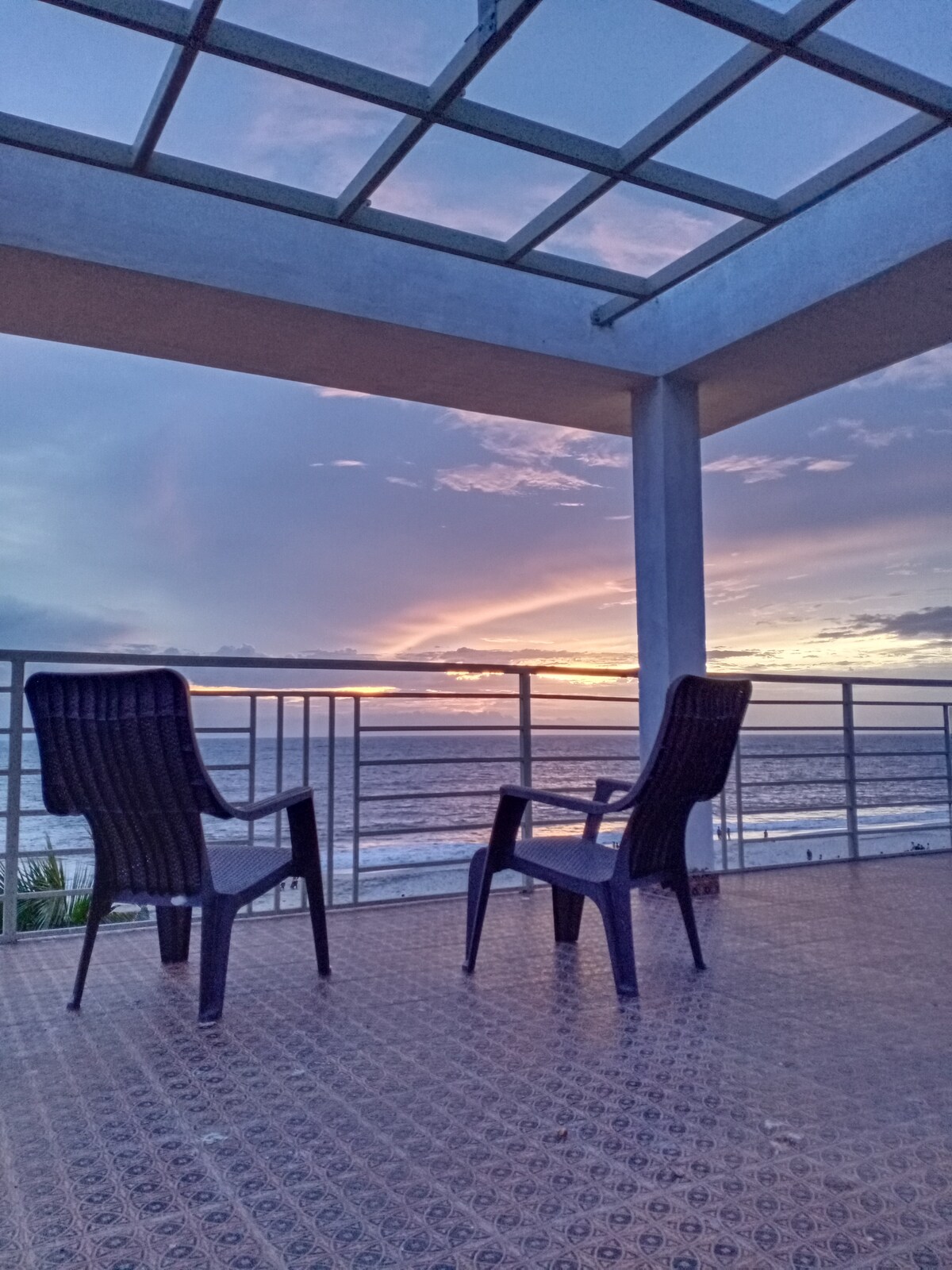 Two black chairs are positioned on a spacious balcony, which offers an expansive view of the sea at sunset. The sky is adorned with hues of purple and orange, creating a serene backdrop. The balcony features a railing that enhances the open view.
