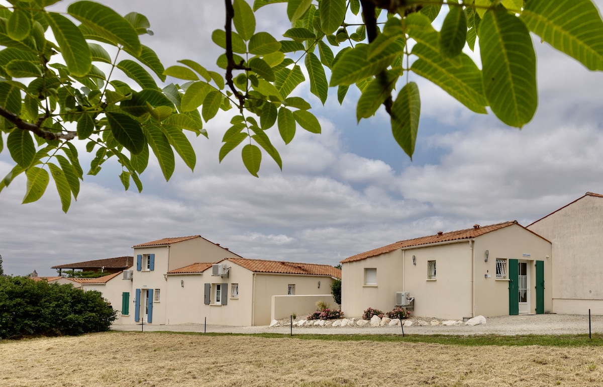 The image presents a view of several independent pavilions within a residence. Each pavilion features a light exterior with tiled roofs and green doors. Surrounding greenery and a grassy area enhance the peaceful and spacious environment.
