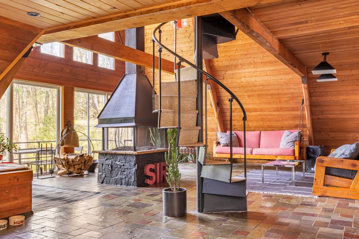 A spacious living area features high wooden ceilings, warm tones, and large windows that bring in natural light. A spiral staircase leads to the upper level. A cozy seating area is arranged around a central wood-burning fireplace, enhancing the inviting atmosphere of the space.