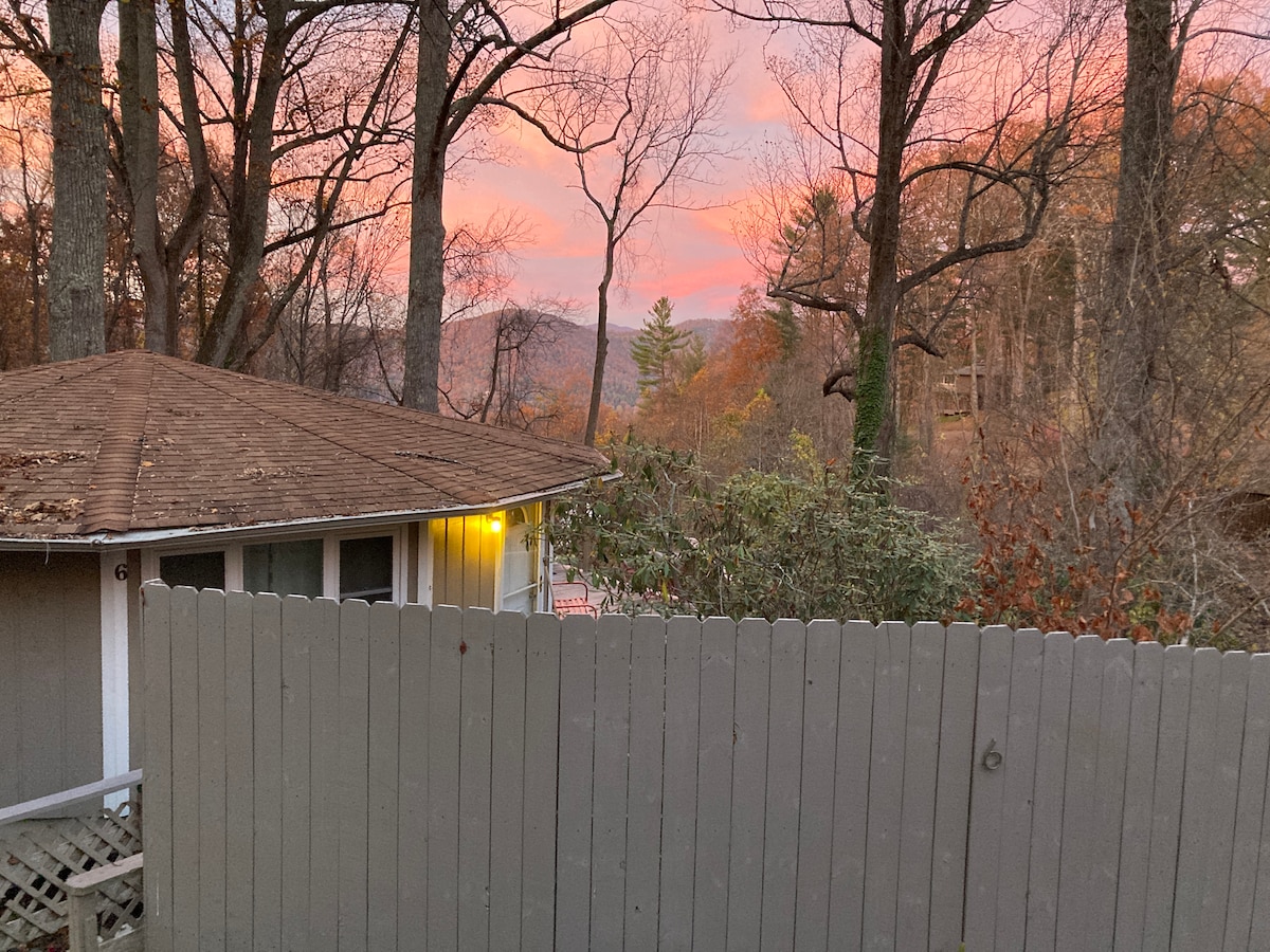 A scenic view captures a round house nestled among trees, with a wooden fence in the foreground. A soft glow from a light illuminates the exterior, while a pastel sky at sunset casts calming colors over the mountainous landscape in the distance.