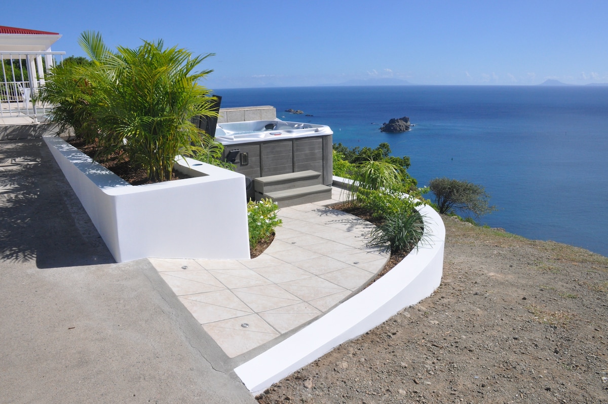 A heated spa is situated on a well-maintained terrace, surrounded by lush greenery. The spa overlooks the expansive Caribbean sea, providing unobstructed views of the horizon. The area features stone tiles and a smooth concrete pathway leading to the edge of the property.
