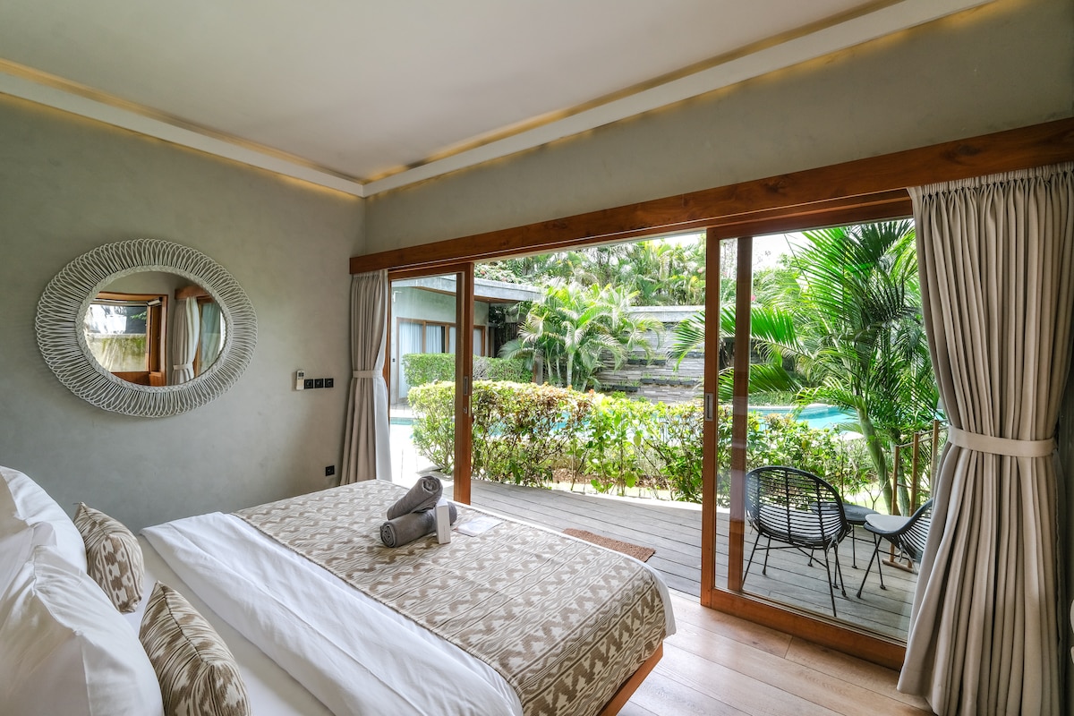 The room features a spacious bed with neutral-toned bedding, complemented by a decorative mirror on the wall. Floor-to-ceiling glass doors open to a private patio overlooking lush greenery and an inviting pool, with a small seating area positioned outside.