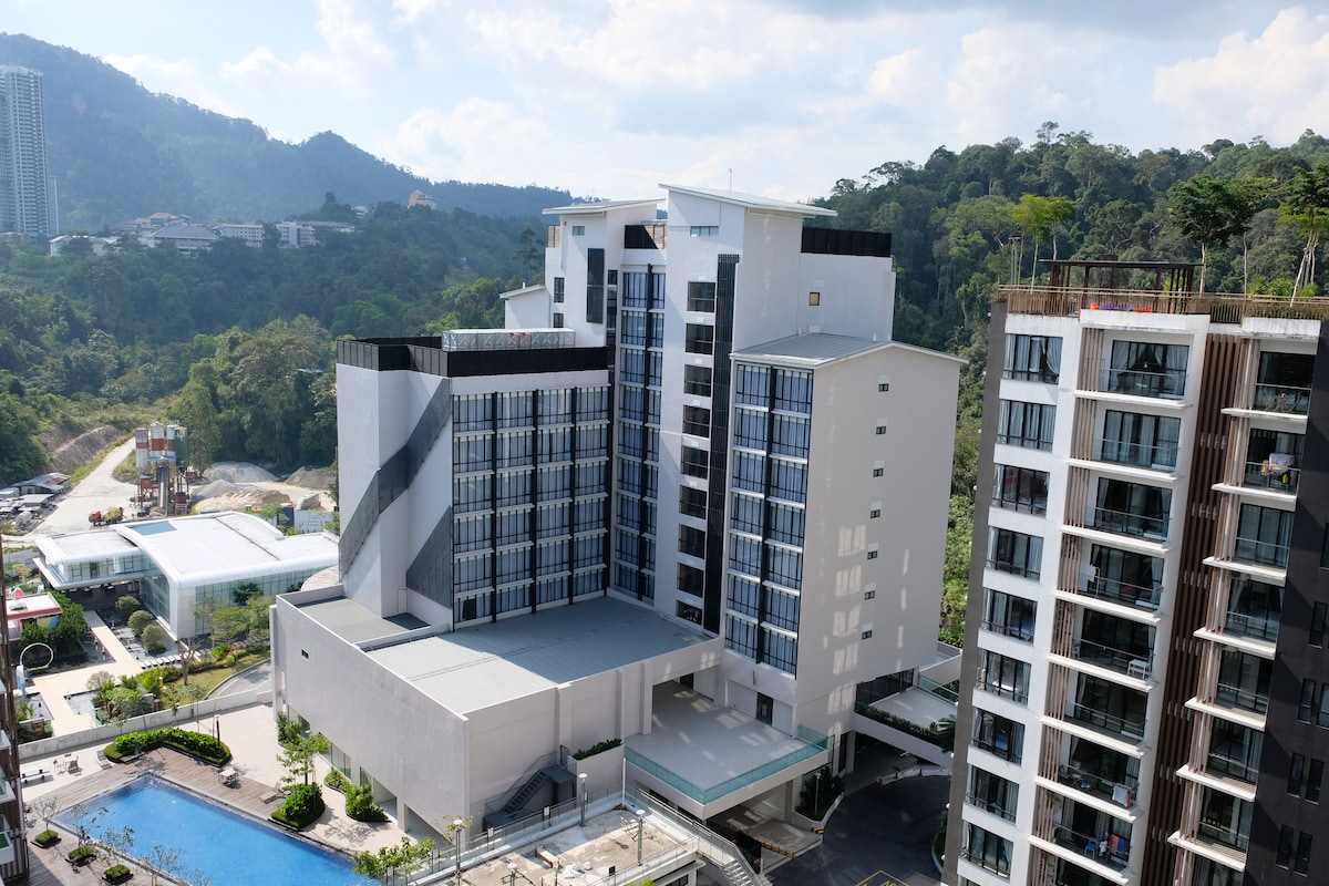 An aerial view captures the modern architecture of a multi-story resort building, bordered by lush greenery and distant mountains. A swimming pool can be seen at the base, while balconies overlook the serene landscape.