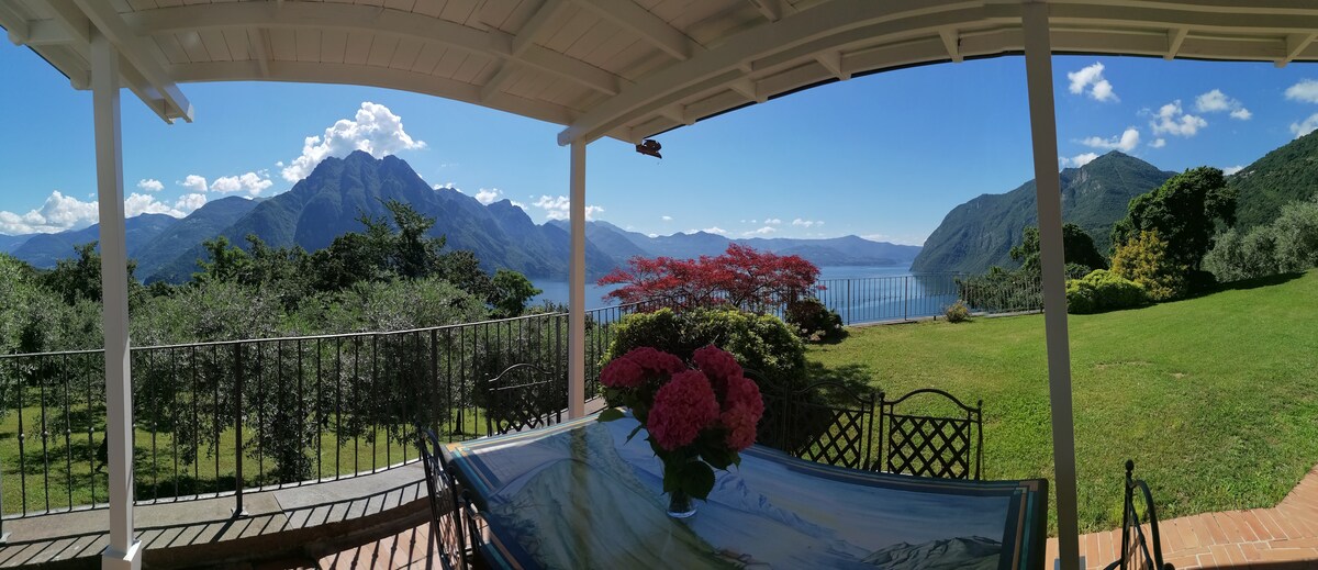 A panoramic view of lush green surroundings and distant mountains is framed by a covered terrace. A table adorned with a floral centerpiece offers a serene space for relaxation, while the lake sparkles under the sunlight, enhancing the tranquil ambiance.