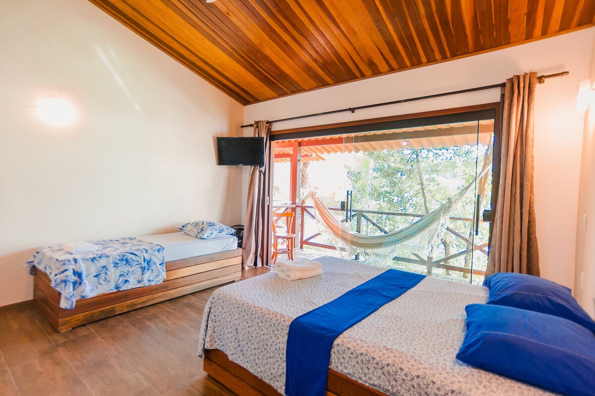 A suite features a spacious bed and an additional single bed. Large glass doors open to a balcony with a hammock, allowing natural light to fill the room. A flat-screen TV is mounted on the wall, and tasteful wood accents enhance the ambiance.