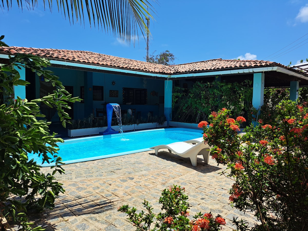 A clear blue swimming pool is surrounded by a stone patio, with a waterfall feature adding to the serene ambiance. Lush greenery and vibrant flowers frame the area, while a sun lounger invites relaxation under the sunny sky.