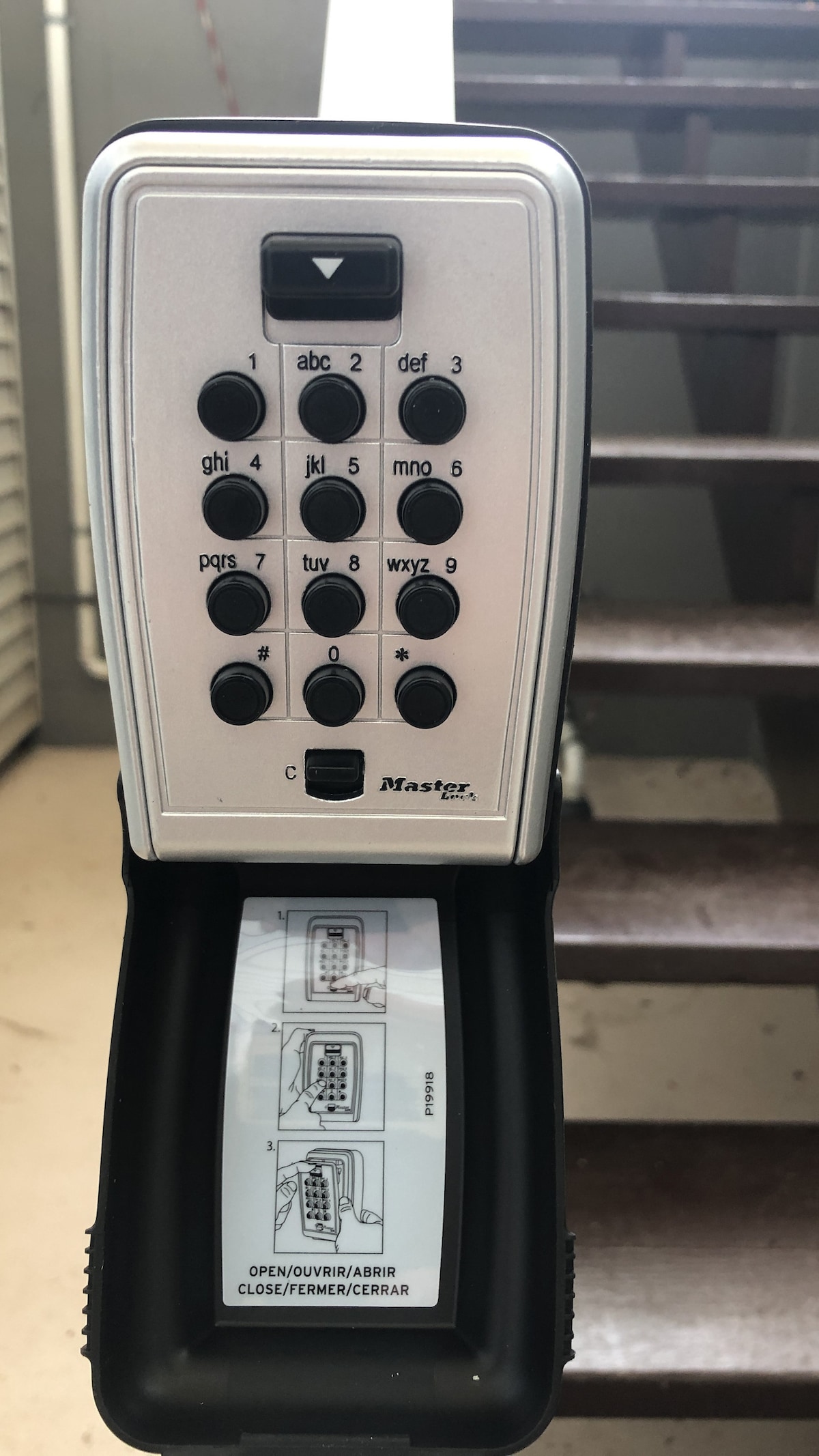 A secured entry lockbox is mounted near a staircase. The lockbox features a keypad with numbered buttons and a compartment storing instruction details. The solid wooden stairs leading to the entrance are visible in the background, providing access to the basement.
