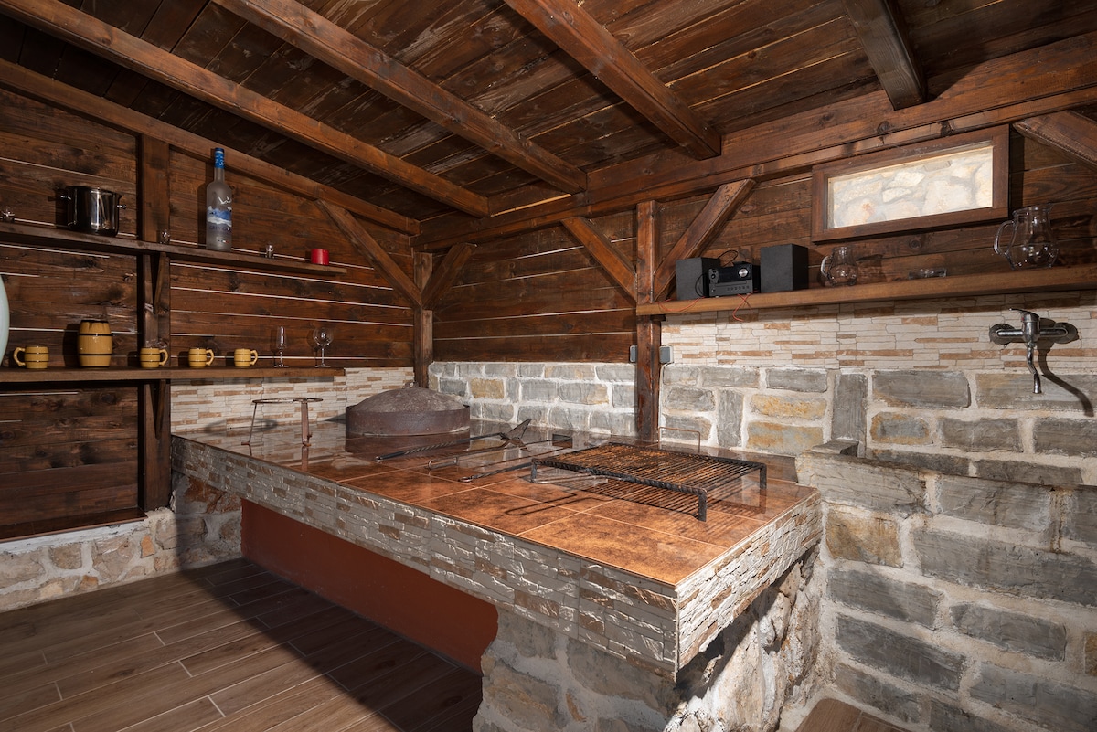 An outdoor kitchen is presented with a rustic wooden structure and stone walls. A large grilling area is centered on a stone countertop, complemented by wooden shelves displaying various utensils and glasses. The warm wood tones create an inviting cooking space.