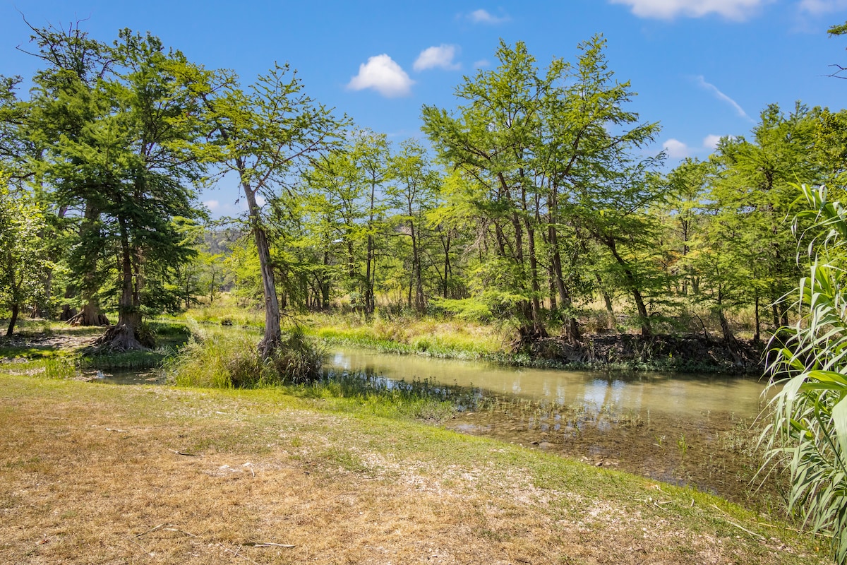 Lush greenery surrounds a tranquil stretch of the Guadalupe River, with dense trees lining the water's edge. The river flows gently, reflecting the clear blue sky above. A mix of grass and gravel forms the shoreline, creating an inviting space for relaxation.