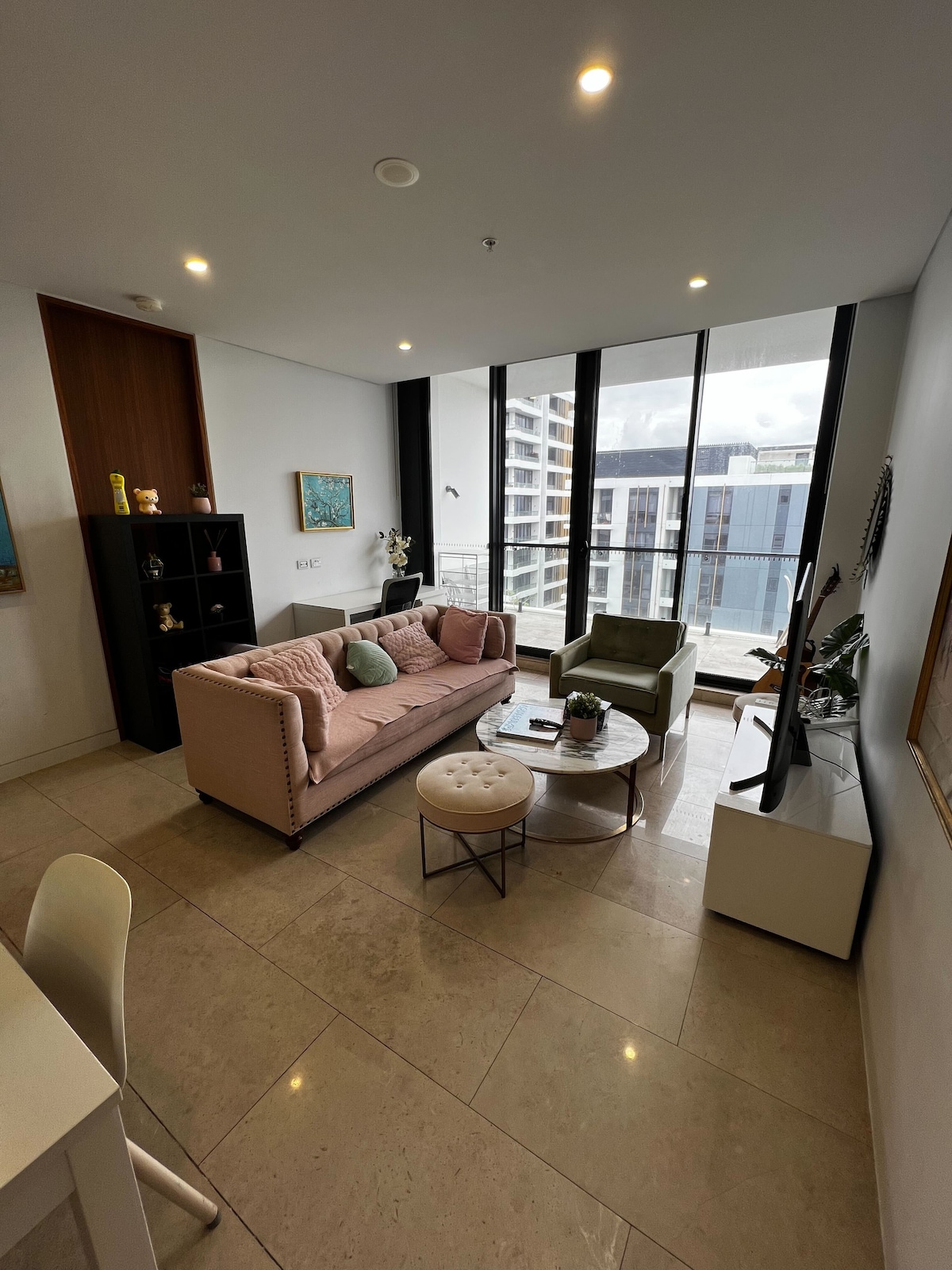 A stylish living area features a pink sofa accompanied by a green armchair, creating a cozy seating arrangement. Large floor-to-ceiling windows offer a view of the outside while allowing natural light to fill the space. A round coffee table sits at the center, enhancing the room's design.
