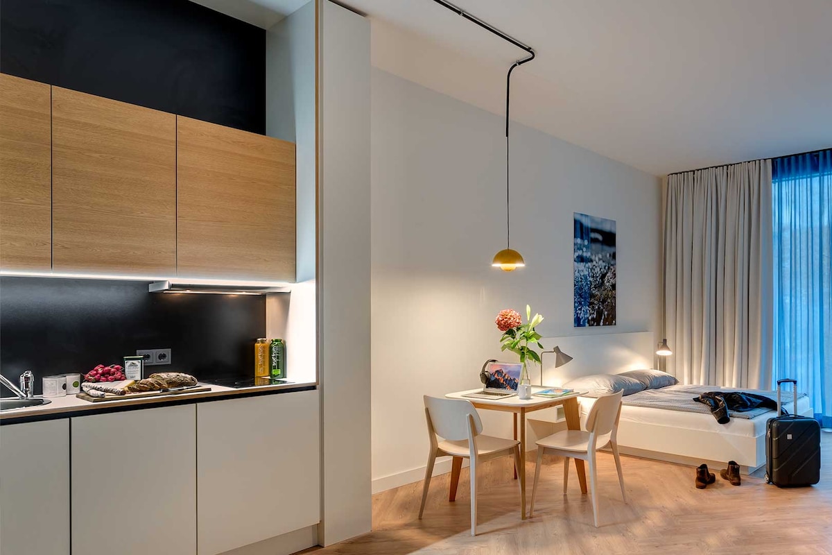 A modern studio apartment is presented, featuring a well-equipped kitchenette with wooden cabinetry. A dining table with two chairs is adjacent to a single bed. A piece of artwork decorates the wall, and soft lighting creates a calm atmosphere. Large windows allow natural light to enter.