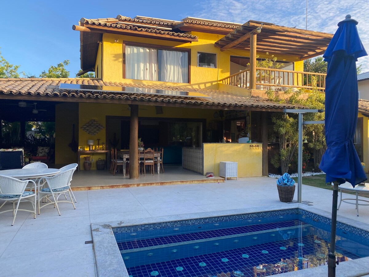 A spacious outdoor area showcases a pool surrounded by a tiled deck. The vibrant yellow house features a large balcony with wooden beams and multiple seating areas. Lush greenery enhances the inviting ambiance, while an umbrella provides shade next to the pool.