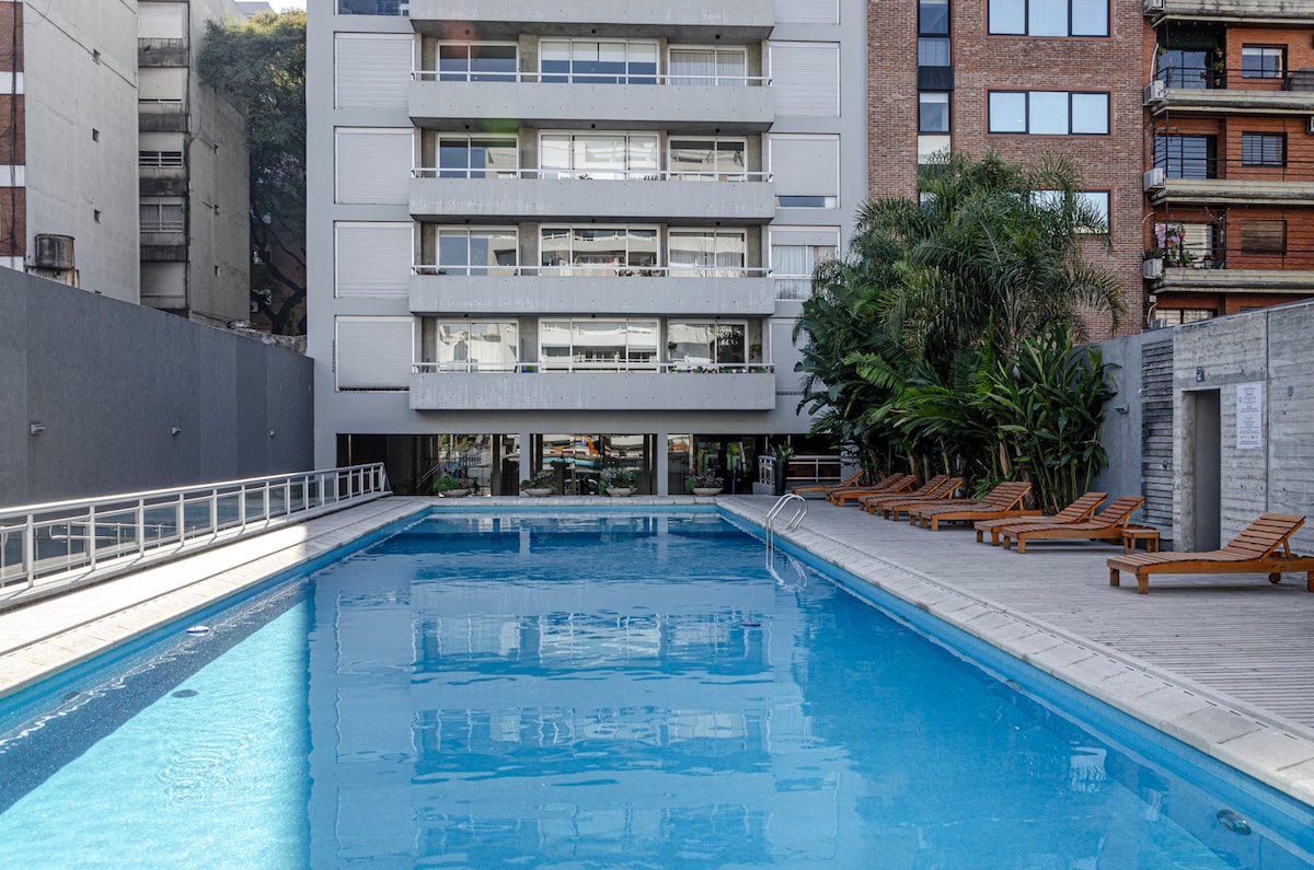 An outdoor pool is framed by a wooden deck and lounge chairs, surrounded by lush greenery. The modern building's façade is visible in the background, enhancing the serene atmosphere of the pool area.
