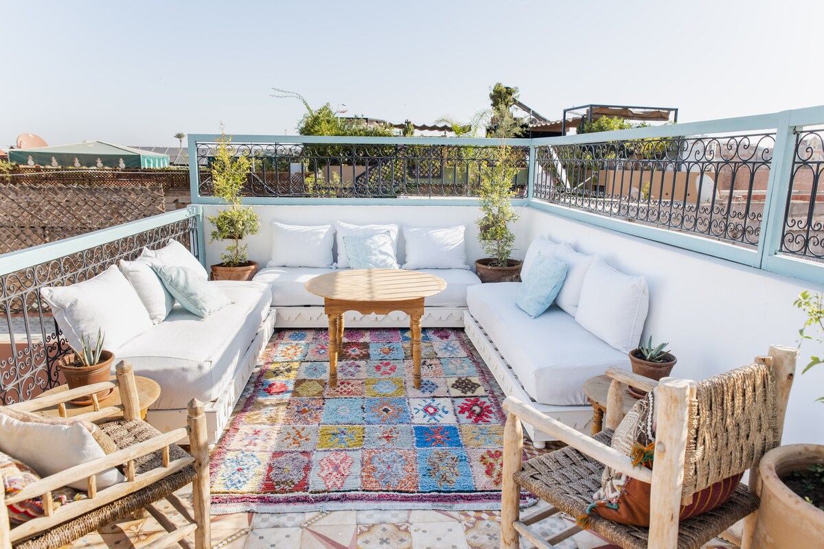 A shared terrace area features comfortable seating with plush white cushions and a colorful patterned rug beneath a wooden table. Potted plants add a touch of greenery, and a clear sky creates a bright atmosphere.