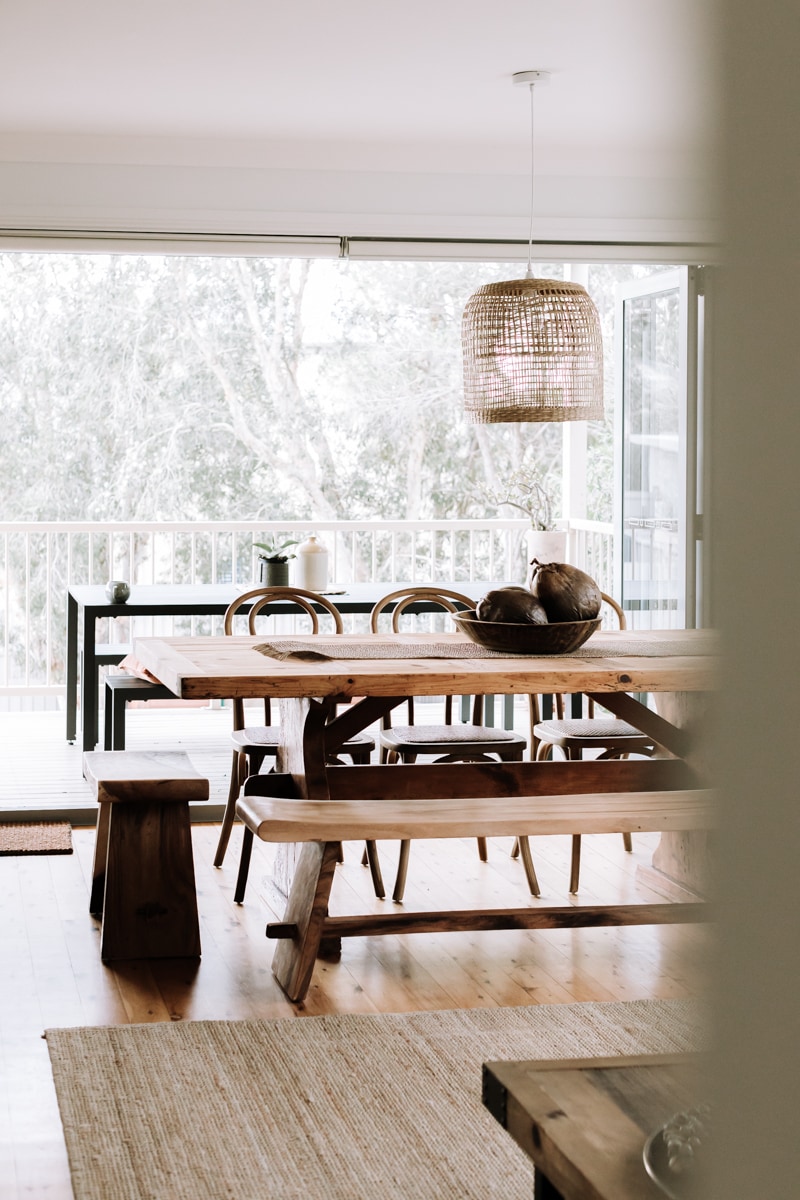 A spacious dining area features a large wooden table surrounded by various seating options. Natural light filters in through large sliding doors, opening to a deck. A woven pendant light hangs above the table, creating a relaxed atmosphere.