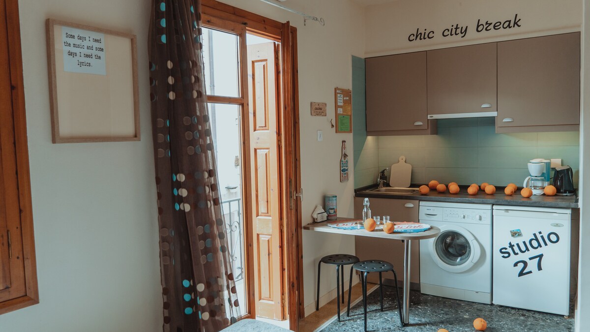 A bright kitchenette is visible, featuring a washing machine and a small dining table set for two. Light-colored cabinetry contrasts with the vibrant scene of fresh oranges arranged along the table. Large wooden windows with shutters illuminate the cozy space, enhancing the urban vibe.