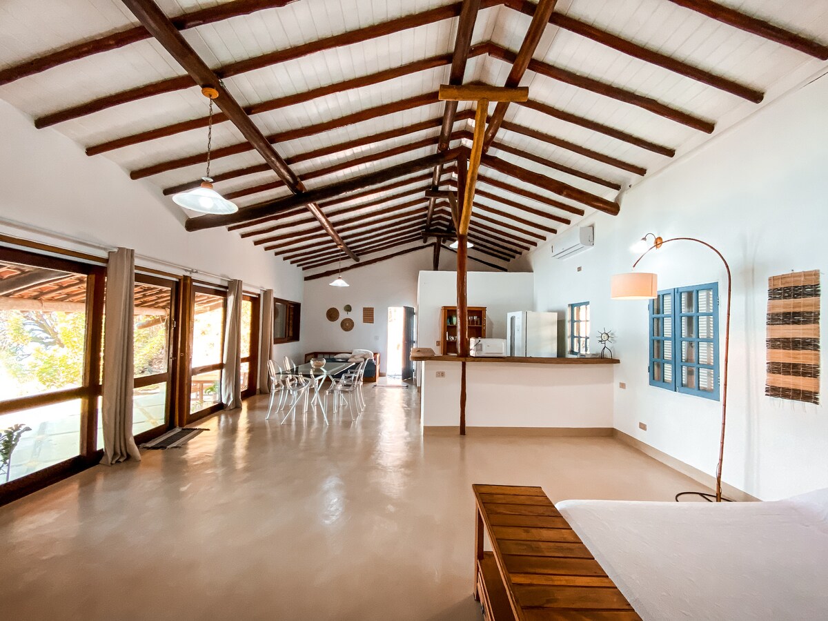 A spacious open-plan living area features high, sloped wooden ceilings and large windows allowing natural light to fill the space. A dining table and an inviting kitchen counter are visible, along with comfortable seating. The warm tones of the wood create a cozy environment.