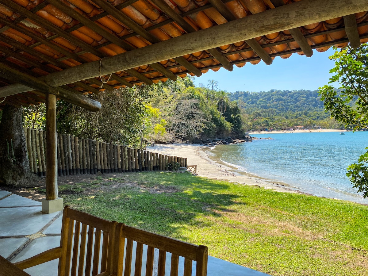 A shaded outdoor seating area overlooks a serene beach and calm blue waters. A gentle curve of the shoreline is visible, framed by lush greenery and distant hills. The rustic wooden structure provides a natural connection to the surroundings.