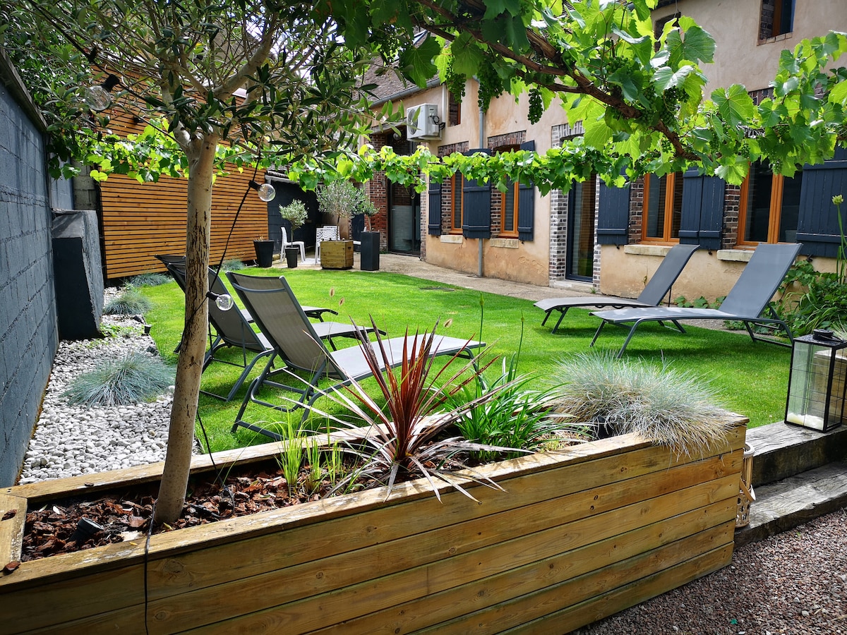 A well-maintained garden features a green lawn surrounded by neatly arranged plants in wooden planters. Two lounge chairs are positioned for relaxation, with decorative landscaping elements visible throughout the space. A small pathway leads to the entrance of the property.