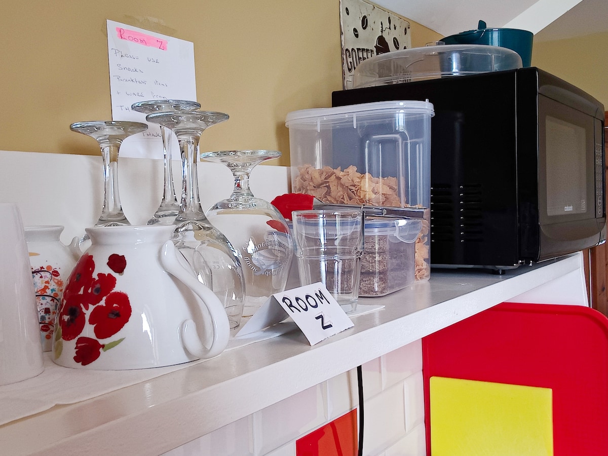 A neatly arranged kitchen shelf displays glassware, including wine glasses and a teapot adorned with a floral design. Clear containers hold breakfast items, while a microwave is positioned at the back. Labelled items suggest organization for guest convenience.