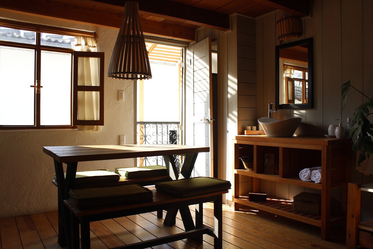 An inviting dining area features a wooden table surrounded by benches adorned with cushions. Natural light filters through the open window and door, casting gentle shadows. A decorative light fixture hangs above, and a wooden console with toiletries is positioned beside a mirror.