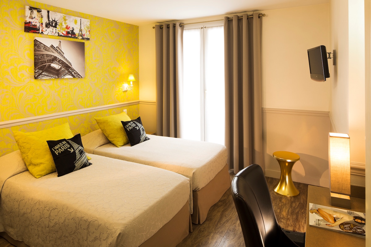 A welcoming guest room features two individual beds with cream-coloured bedding, accented by yellow decorative pillows. A wall displays an art piece of the Eiffel Tower. Natural light enters through the window, complemented by grey curtains. A flat-screen television, desk, and lamp are also present.