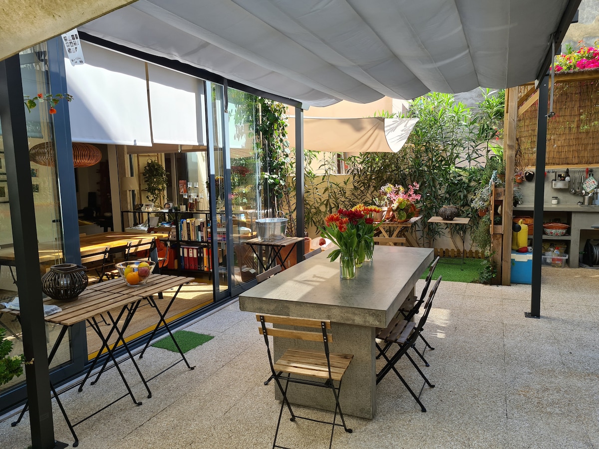 A spacious garden area features a large concrete dining table surrounded by wooden chairs. Colorful flowers in a vase sit atop the table. Large windows and an outdoor kitchen area are visible, with greenery and decorative plants creating a refreshing ambiance.