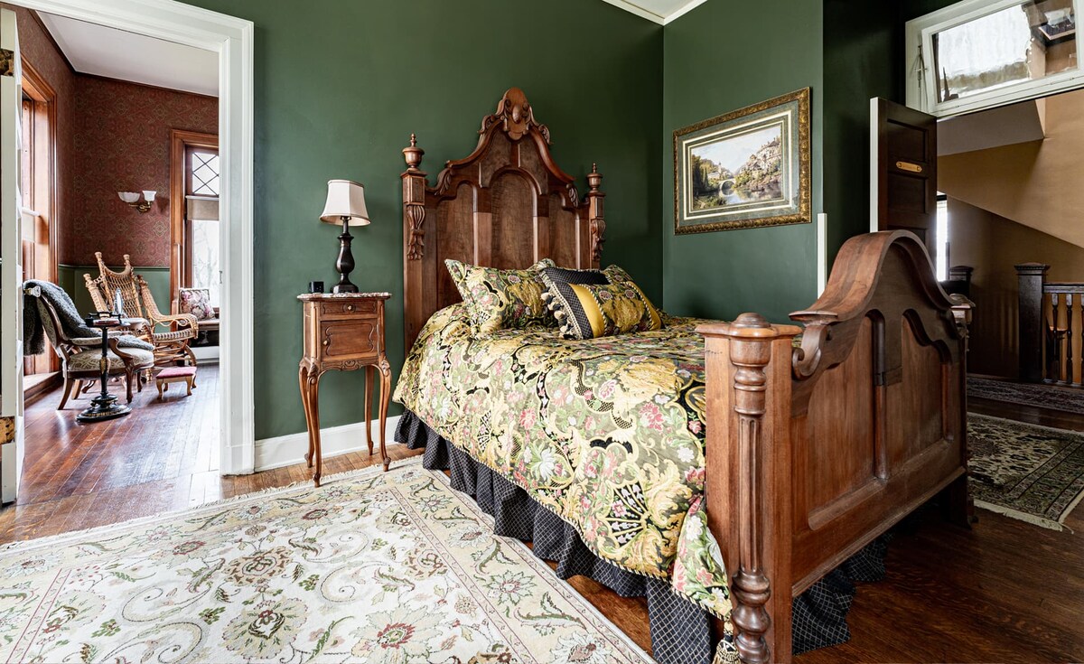The bedroom features a high back burl-wood-posted queen-size bed with patterned bedding and an ornate headboard. A bedside lamp provides soft lighting. Rich green walls and a patterned area rug complement the classic decor, while a doorway leads to the adjoining sitting room.